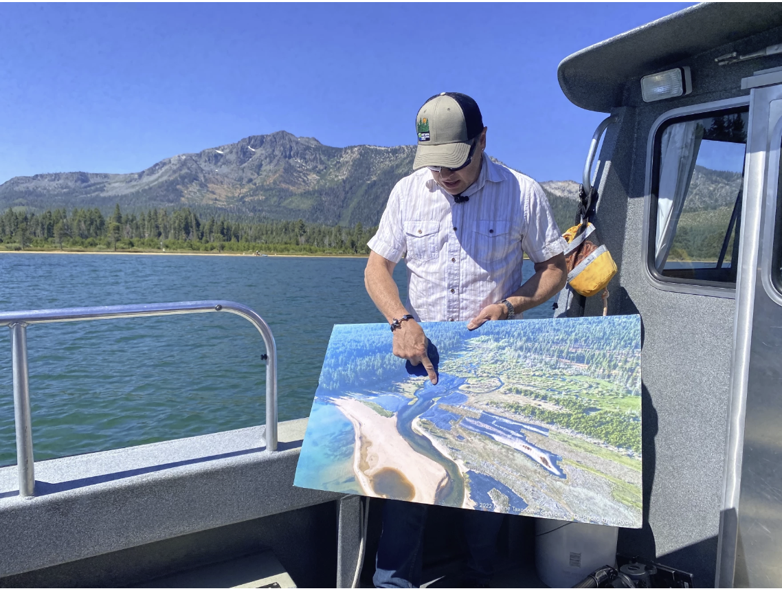 Dennis Zabaglo stands on a boat in front of the Taylor and Tallac creek marsh, pointing to a poster detailing the restoration project on July 29, 2024. Credit: Sydney Peerman / KUNR Public Radio